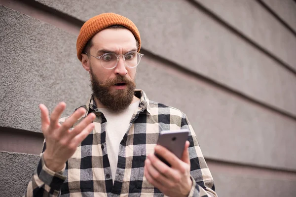 Σοκαρισμένος Άντρας Γένια Στέκεται Smartphone Κοντά Στο Γκρίζο Τοίχο Στο — Φωτογραφία Αρχείου