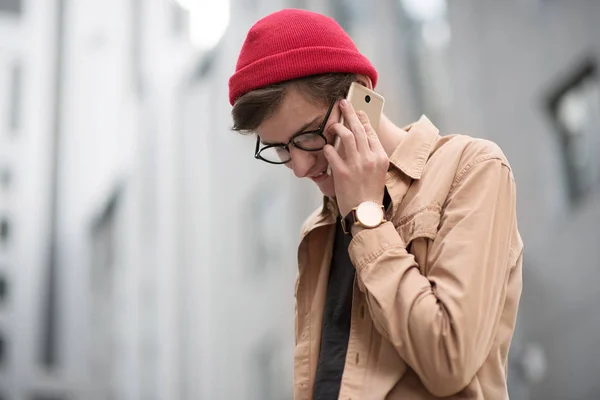 young teenage boy, a hipster, walks the metropolis city in a red hat and a stylish shirt and looks through something in his phone listening to music and having a rest with a good mood