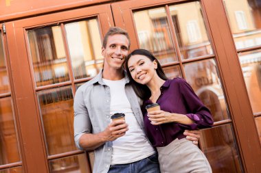 tek kullanımlık kahve fincanları tutan ve kameraya bakarak neşeli genç çift