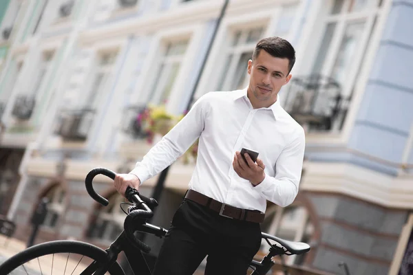 handsome man in shirt standing with bicycle and using smartphone in city
