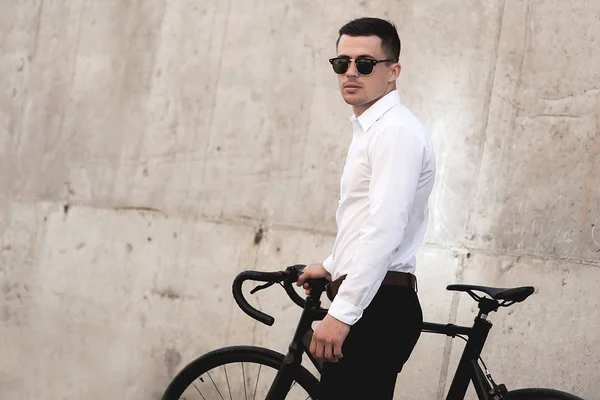 handsome man in sunglasses standing with bike near wall on street