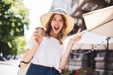 Gülümseyen şık kadın sokakta yürüyüş ve sokak kafe yakınında yalnız iyi duygular ile fincan paket bazı kahve içme iyi bir yaz ruh hali sahip. Yürüyen mutlu bir kız.