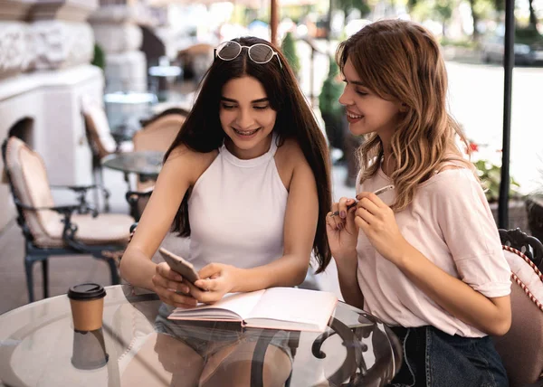 Two pretty young women sitting in street cafe together, drinking coffee, laughing and looking in phone, reading some messages and smiling. They having fun and enjoy summer