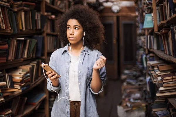 Smart intelligent woman standing near long wall full of bookshelf with different books. She is holding a phone in hand and looking to the books. She listening e-books online and searching offline