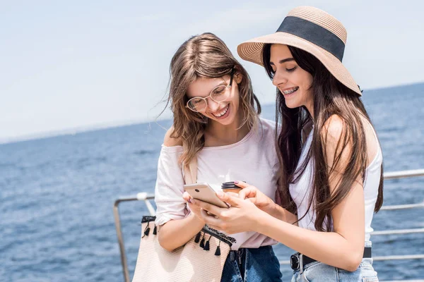 漂亮的年轻女孩与手机智能手机的乐趣 显示照片从度假的海洋和微笑在一起 他们是在阳光明媚的国家度假最好的朋友 — 图库照片