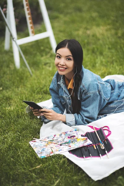 Young woman sitting with arts painting oil pictures and listening music on mobile on grass. Woman studying outdoors with copy-book. Education, inspiration and remote working concept.