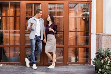 tek kullanımlık kahve fincanları tutan ve kafenin girişinde duran neşeli genç çift