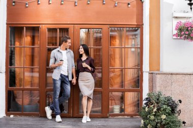 tek kullanımlık kahve fincanları tutan ve kafenin girişinde duran neşeli genç çift