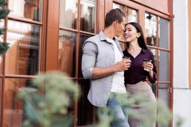 tek kullanımlık kahve fincanları tutan neşeli genç çift ve kafegirişinde yakın öpmek için gidiyor