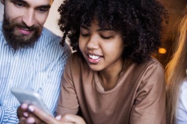 Kıvırcık saçlı şık hipster genç çift modern iç kafede kapalı oturan akıllı telefon bakarak ve gülümseyerek