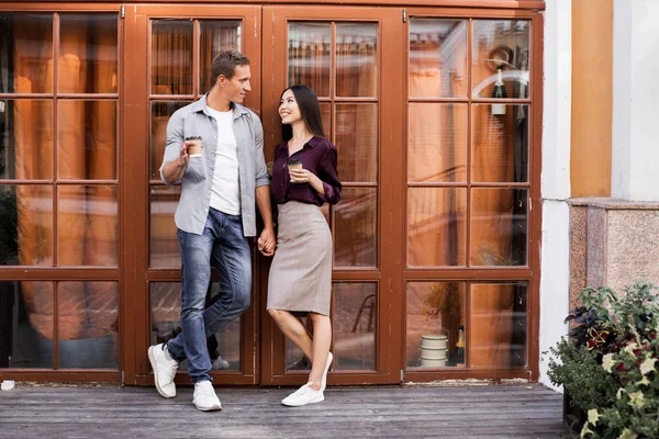 tek kullanımlık kahve fincanları tutan ve kafenin girişinde duran neşeli genç çift