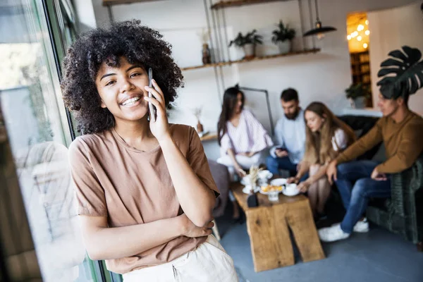 African American young women holding cellphone in hands near the window with calm emotions. A lit of people on background