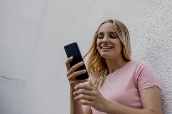 Χαμογελαστή Ελκυστική Ξανθιά Γυναίκα Χρησιμοποιώντας Smartphone Κοντά Στο Γκρίζο Τοίχο — Φωτογραφία Αρχείου