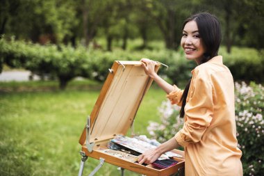 Bir genç mutlu moda Asyalı kadın şehir parkı açık doğada sıcak güneşli bir günde bir şelon bir resim boyama başlıyor. Sanat terapisi, dinlenmek ve hobi kavramı, tatil dinlenme günü