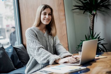 Pretty konsantre kadın freelancer yayın için fikirler dizüstü bilgisayar ile masada oturan, netbook ve ücretsiz kablosuz bağlantı kullanarak kahve dükkanında uzaktan çalışan yetenekli hipster kız