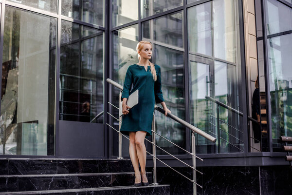 Attractive business woman coming out from modern business center with computer wearing official style after presentation on work meeting, urban space background. Business and finance