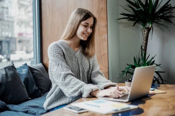 Pretty konsantre kadın freelancer yayın için fikirler dizüstü bilgisayar ile masada oturan, netbook ve ücretsiz kablosuz bağlantı kullanarak kahve dükkanında uzaktan çalışan yetenekli hipster kız