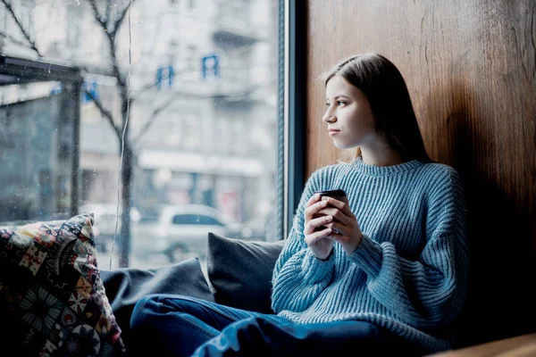 Beautiful pretty woman with serious face drinking coffee near window in cafe near her home. Sad mood, inspiration mood. Waiting for some news or for someone