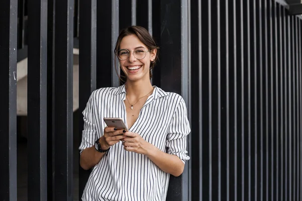 Şehir caddesinde şehir duvarının yanında akıllı telefon tutan çekici mutlu İspanyol kadının portresi. İş konsepti, yaratıcı işçi, break, yürüyüş sokak, resmi giysiler, şık ve başarılı