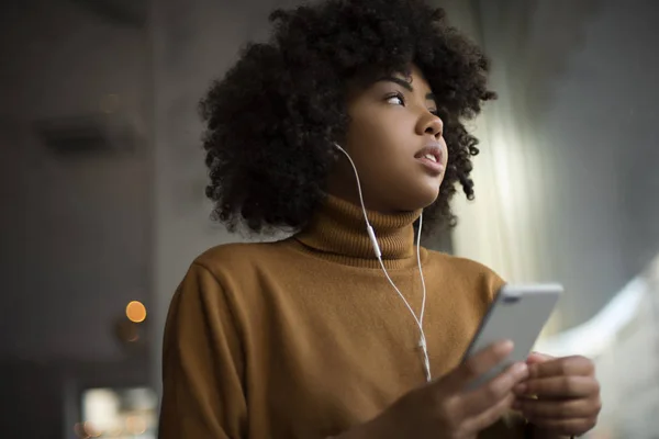 Afro-Amerikan kadın beyaz kulaklıklar, bir hafta sonu günü romantik bir ruh hali ile telefonunda müzik dinlerken bir şehir kahve dükkanının penceresinden bakıyor. Rahat bir tarzda giyinmiş 