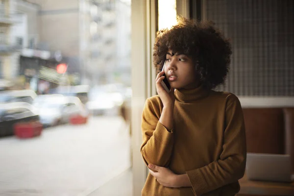 Genç bir Afro-Amerikan stajyer kadın müşterileri ile telefonda konuşurken modern bir loft tarzı ofiste pencerenin yanında duruyor ve akşam şehirde pencereden dışarı bakar. Çevrimiçi uzaktan çalışma