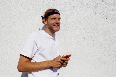 Şık sakallı zencefil kızıl saçlı adam hipster elinde cep telefonu ile beyaz duvar arka plan üzerinde izole yüzünde gülümseme ile uzağa bakarak, mutlu genç adam modern dijital teknolojileri kullanarak. 