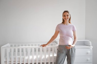 Çocuk odasındaki arenanın yakınında gülümseyen genç ve mutlu hamile bir kadın çerçevede gülümseyen ve elini karnının üstünde tutan pembe tişörtlü bir anne olmaya hazırlanıyor.