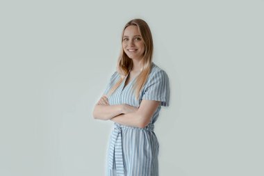 Genç, sıradan bir kadın, kameraya bakıyor, kollarını kavuşturmuş, gri arka planda izole edilmiş.