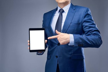 Mavi takım elbiseli bir iş adamı gri-mavi arka planda bir tablet tutuyor ve parmağıyla tableti işaret ediyor. Ürün veya hizmet sunumu, satış veya performans kavramı.