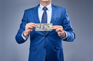 Gri-mavi arka planda mavi takım elbiseli bir işadamı önünde yüz dolarlık banknot tutuyor. Finansal işlemlere katılarak gelir elde etme kavramı.