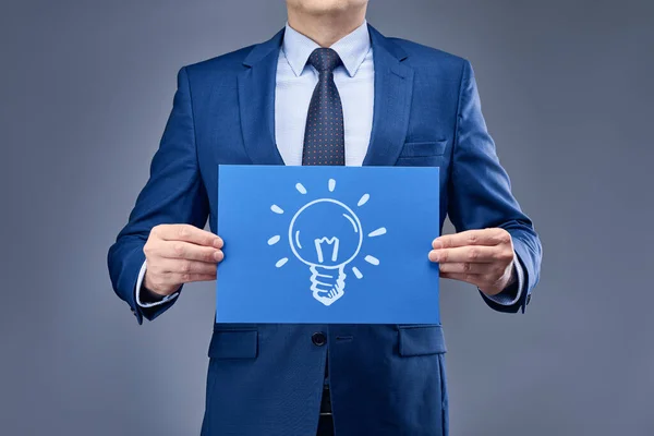 A businessman in a blue suit on a gray-blue background holds a sheet of blue paper in front of him with the image of the idea sign. Concept of product or service presentation, sales, or performance.