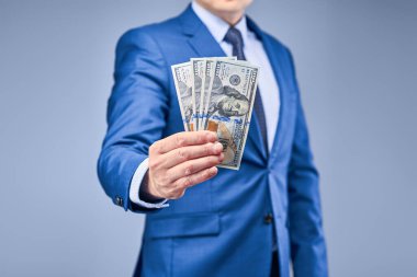 Gri-mavi arka planda mavi takım elbiseli bir işadamı önünde dört yüz dolarlık banknotlar tutuyor. Elle yakından çek. Finansal işlemlere katılarak gelir elde etme kavramı.