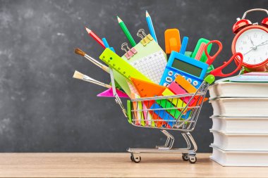 Okul için çeşitli ofis malzemeleri bir el arabası, ders kitapları ve bir çalar saat, ahşap bir masanın üzerinde bir tahtaya karşı. Okul hazırlığı kavramı, yeni bir okul yılının başlangıcı..