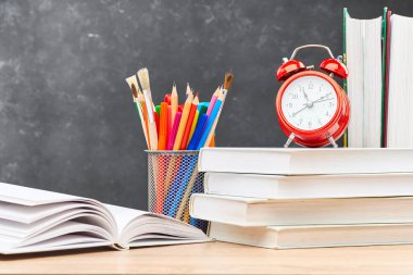 Bir yığın ders kitabı, açık bir ders kitabı, stantta duran kalemler ve tahta bir masanın üzerindeki çalar saat bir tahtaya karşı. Yeni akademik yılın başlangıcına hazırlık kavramı