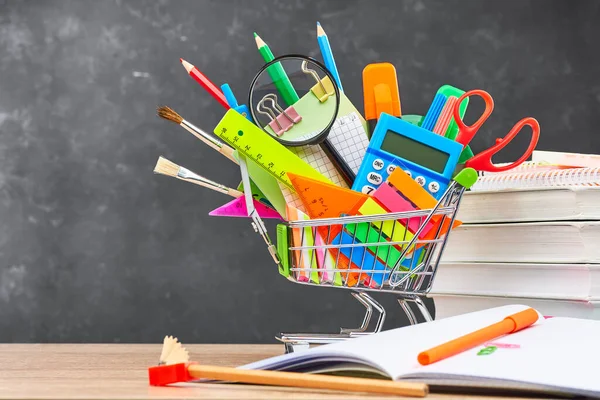 Various office supplies for school in a cart, textbooks and an open notebook, on a wooden table against a blackboard. The concept of preparing for school, the beginning of a new school year.