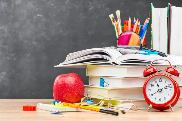 Textbooks, pencils in a stand, a magnifying glass, an alarm clock,and stationery on a wooden table against a blackboard. Concept of preparation for the beginning of the new academic year.