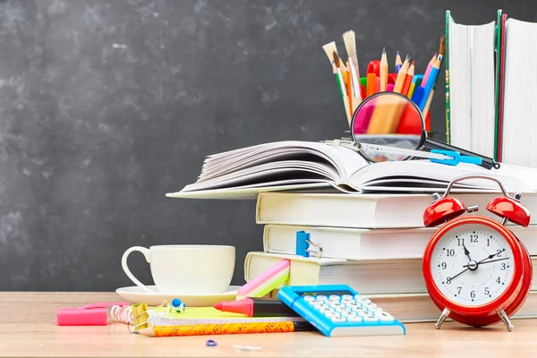 Textbooks, pencils in a stand, a magnifying glass, an alarm clock, a calculator and stationery on a wooden table against a blackboard. Concept of preparation for the beginning of the new academic year