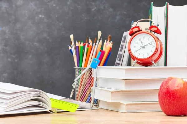 A stack of textbooks, an open textbook, pencils in a stand, an alarm clock,and a red Apple on a wooden table against a blackboard. Concept of preparation for the beginning of the new academic year