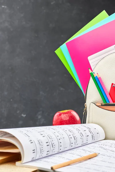A school bag with colored notebooks and pencils, a notebook with notes and a red Apple on a wooden table against a black Board. Concept of preparation for the beginning of the new academic year