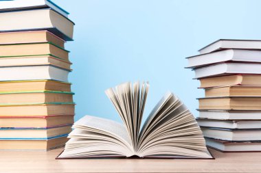 Açık bir kitap ve iki kitap yığını, açık mavi bir duvara karşı ahşap bir masanın üzerinde duruyor.