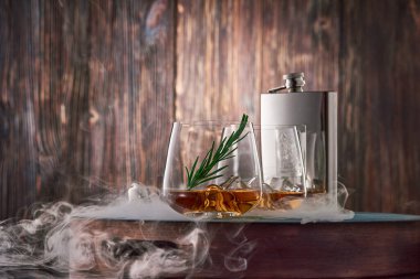 İki kristal bardak buzlu viski ve bir tutam biberiye ve dumanla kaplanmış metal bir matara eski ahşap bir duvara karşı tepside duruyor. Alçak tuş.