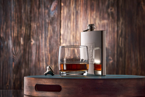 A round glass of whiskey and a metal flask stand on a tray against an old wooden wall. Low key.