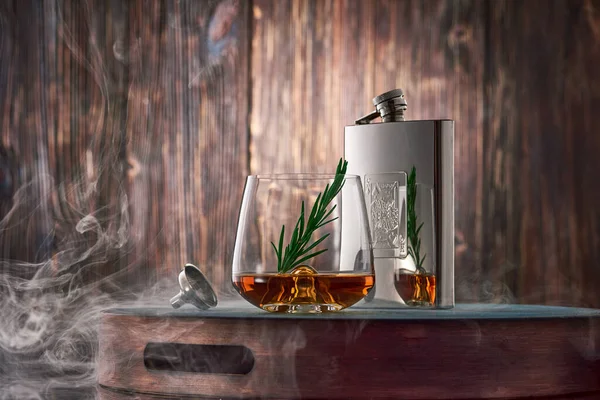 A crystal glass of whiskey with a sprig of rosemary and a metal flask, shrouded in clouds of smoke, stand on a tray against an old wooden wall. Low key.