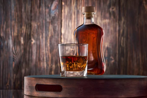 岩の上にあるウイスキーの正方形のガラスと古い木製の壁に対するトレイの上にウィスキーの完全なボトルが立っています 鍵が低い — ストック写真