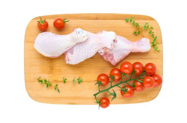 Tahta bir kesim tahtasının üzerinde üç tane taze tavuk budu ve beyaz arka planda izole edilmiş sebze ve otlar. Yukarıdan görünüşü.