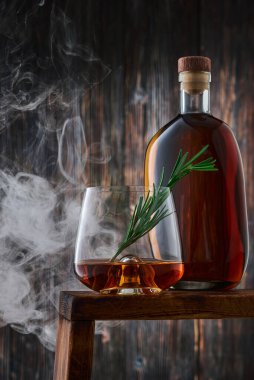 Bir kadeh viski, bir tutam biberiye ve tezgahın üzerinde dumanlı bir şişe viski, eski ahşap duvara karşı. Alçak tuş, dikey.