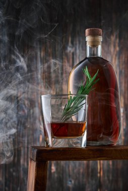 Bir yudum viski ve bir tutam biberiye ile kare bir bardak ve tezgahın üzerinde eski ahşap duvara karşı dumanlı bir şişe viski. Alçak tuş, dikey.