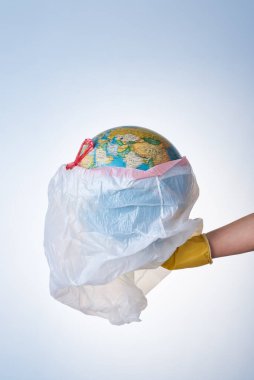 Dünyayı plastik atıklardan, çevre kirliliğiyle mücadele etme kavramı, sarı lastik eldivenli bir elin hafif arka planda küresi olan beyaz bir çöp torbasını tutması. Poşet yok.