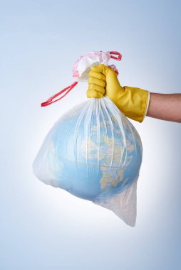 Dünyayı plastik atıklardan, çevre kirliliğiyle mücadele etme kavramı, sarı lastik eldivenli bir elin hafif arka planda küresi olan beyaz bir çöp torbasını tutması. Poşet yok.