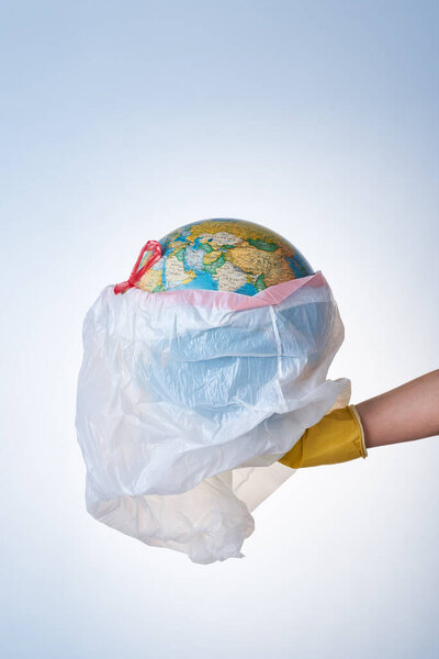 Концепция защиты мира от пластиковых отходов, борьбы с загрязнением окружающей среды, рука в желтой резиновой перчатке держит белый мусорный мешок с глобусом на светлом фоне. Без пластиковых пакетов
